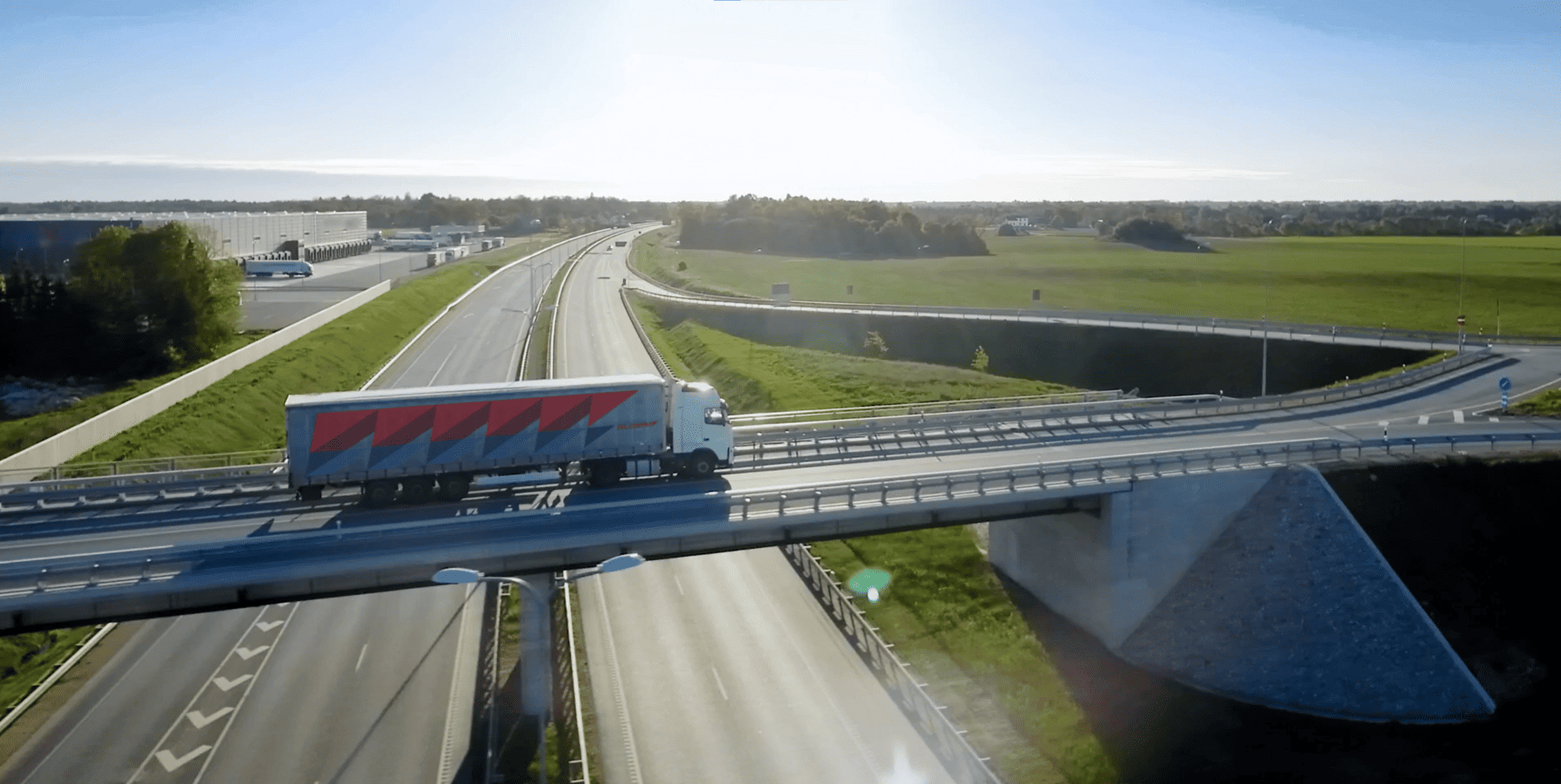 A white truck with red lettering drives over a bridge. Under the clear sky, highways and green fields can be seen.