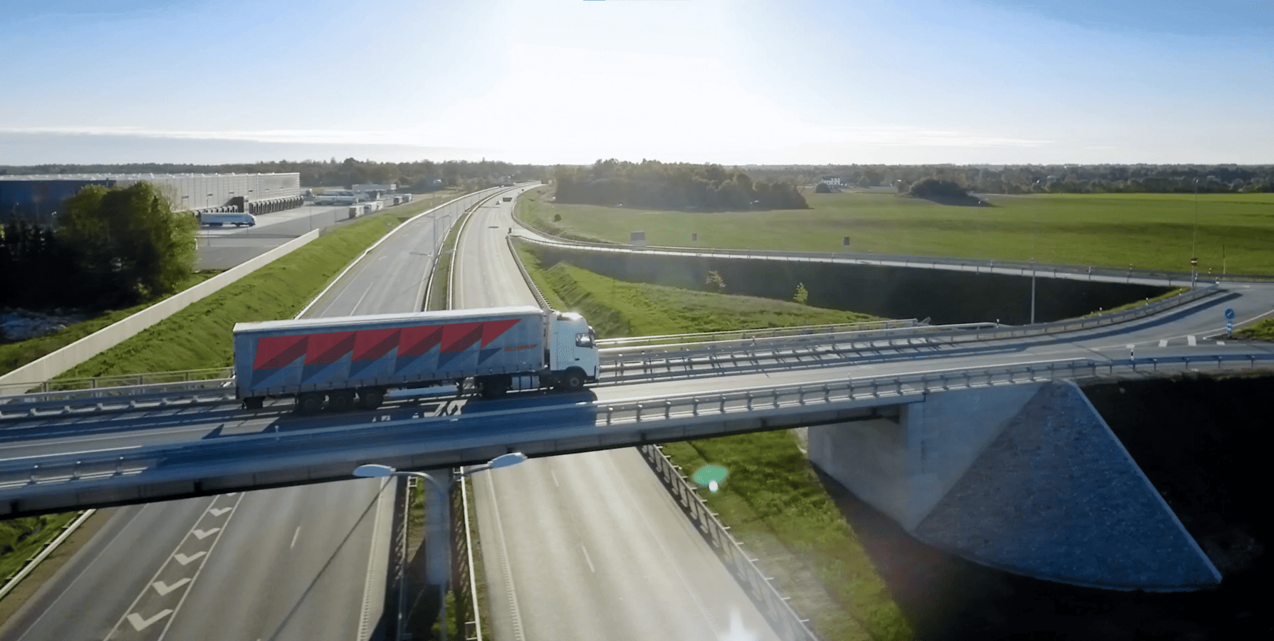 Ein weißer Lkw mit roter Aufschrift fährt über eine Brücke. Unter dem klaren Himmel sind Autobahnen und grüne Felder zu sehen.