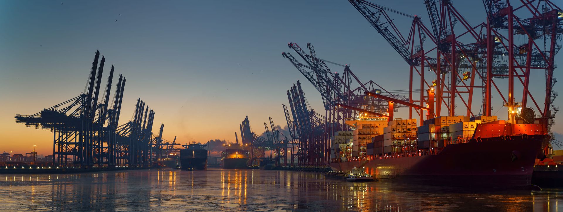 Crane silhouettes and container ships illuminated at dusk in a busy harbor and reflected on the still water.