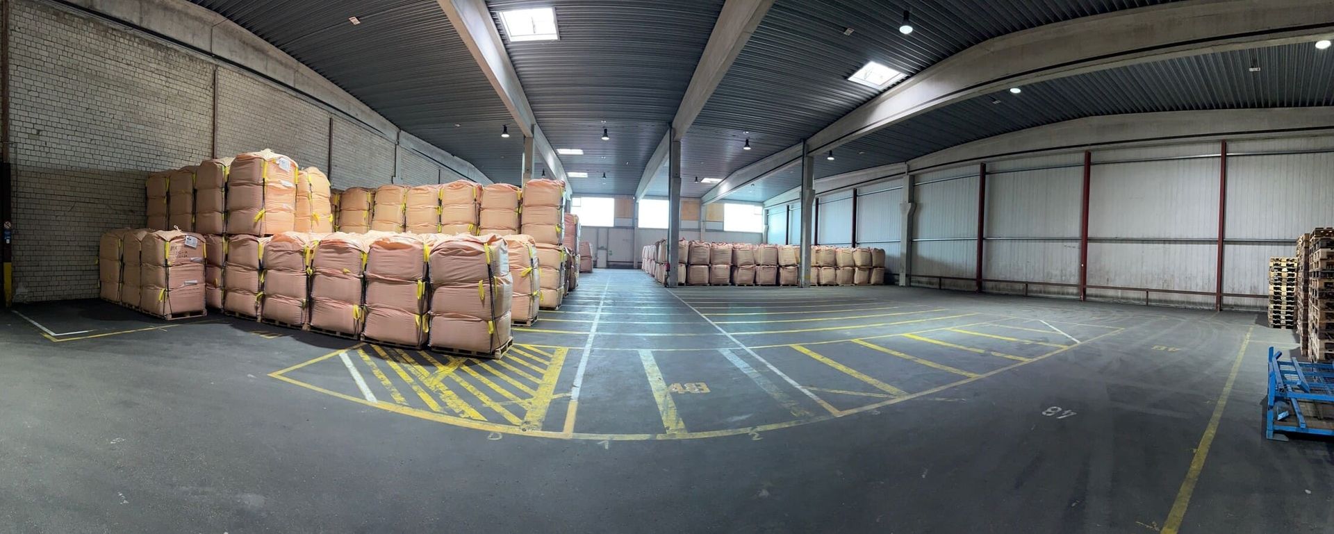 Das Innere eines geräumigen Lagers, gefüllt mit großen, gestapelten und mit gelben Bändern gesicherten Ballen, neben markierten Parklinien.