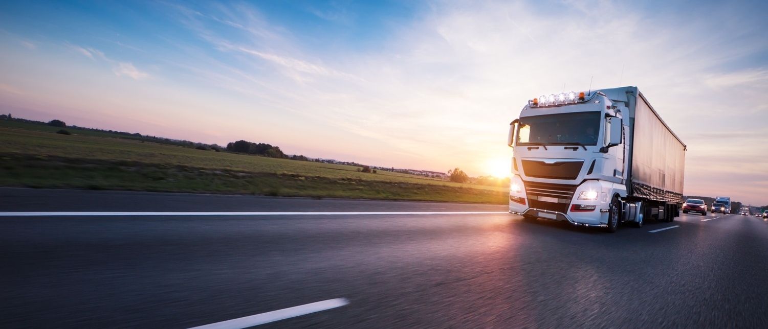 Ein weißer Lastwagen fährt bei Sonnenuntergang auf einer Autobahn, umgeben von grünen Feldern und einem klaren Himmel, mit anderen Fahrzeugen im Hintergrund.