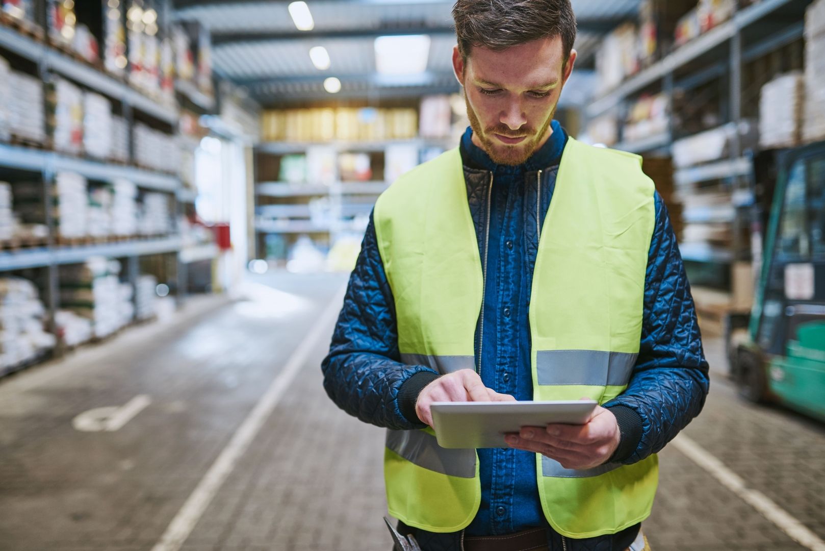 A person in a bright yellow safety vest uses a tablet in a well-organized warehouse with supplies stacked on shelves.